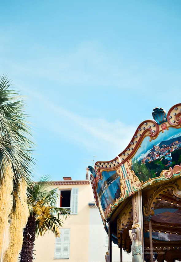 Details from the historic district of a southern France town, showing a palm tree and a carousel in the foreground. Pastel colored house fronts are visible in the background.